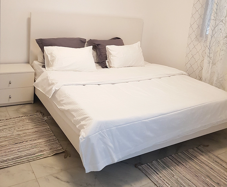 Chambre King Size : grand lit blanc aux coussins gris, tête de lit capitonnée et tapis tissé